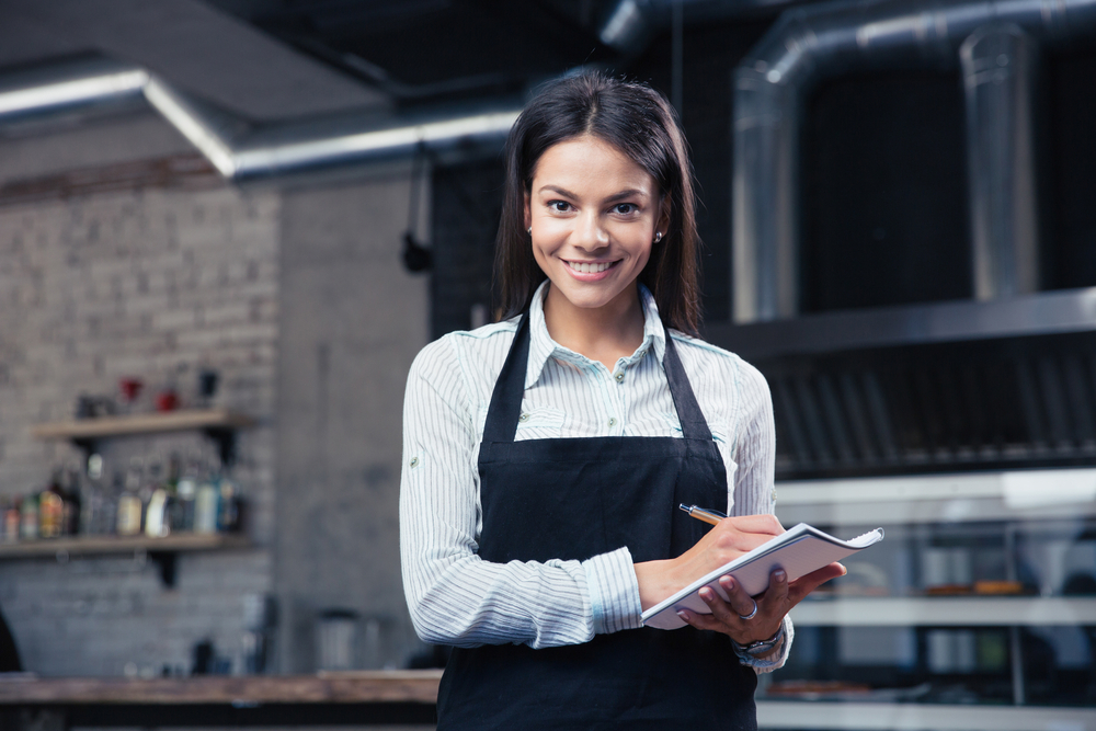 Happy charming female waiter in apron writing order and looking at camera